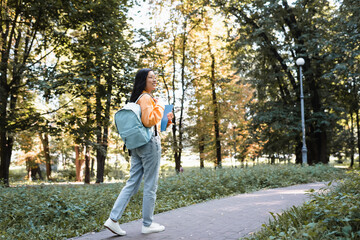 Fototapeta premium smiling asian woman in jeans walking in park with backpack and notebooks