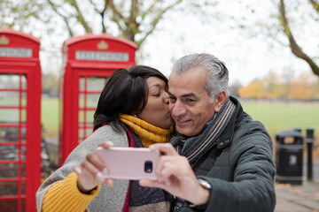 Smiling senior couple taking selfie in autumn park in front of red telephone booths