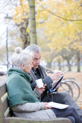 Senior couple reading newspaper and drinking coffee on bench in autumn park