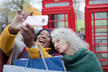 Obraz premium Senior women friends with shopping bags taking selfie in autumn park in front of red telephone booths