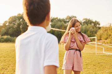 Boy with girl having fun by using tin can telephone while standing outdoors on the field
