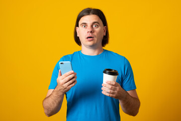 Surprised handsome man uses mobile phone for online communication and surfing internet holds takeaway coffee being in good mood dressed casually