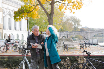 Senior couple with bicycles traveling, looking at guidebook along autumn river