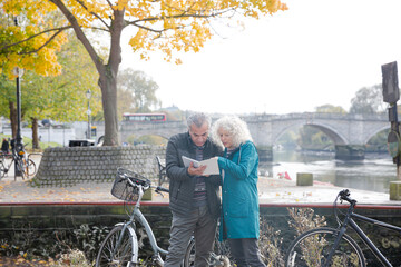 Senior couple with bicycles traveling, looking at guidebook along autumn river