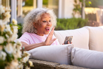 Senior Retired Woman Sitting Outside In Garden At Home Making Video Call On Mobile Phone