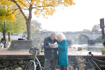 Senior couple with bicycles traveling, looking at guidebook along autumn river
