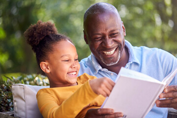 Grandfather Sitting Outdoors With Granddaughter At Home Reading Book Together