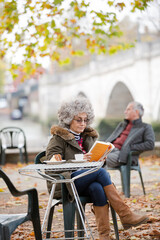 Active senior woman reading book, enjoying cake and coffee at autumn park cafe