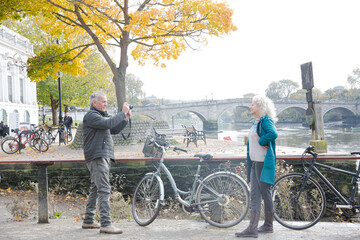 Senior man photographing wife with bicycle in autumn city