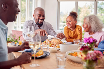 Father Carving As Multi Generation Family Sit Around Table At Home And Enjoy Eating Meal