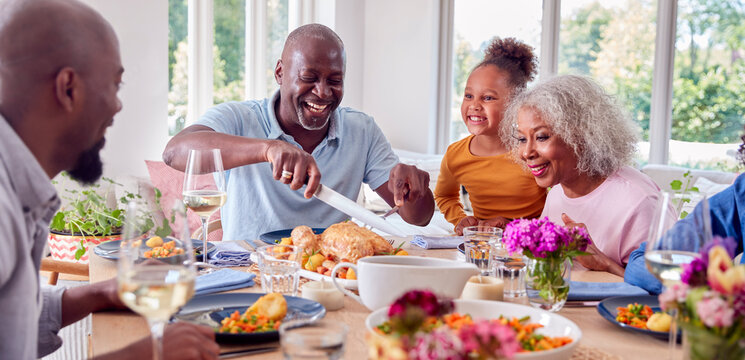 Grandfather Carving As Multi Generation Family Sit Around Table At Home And Enjoy Eating Meal