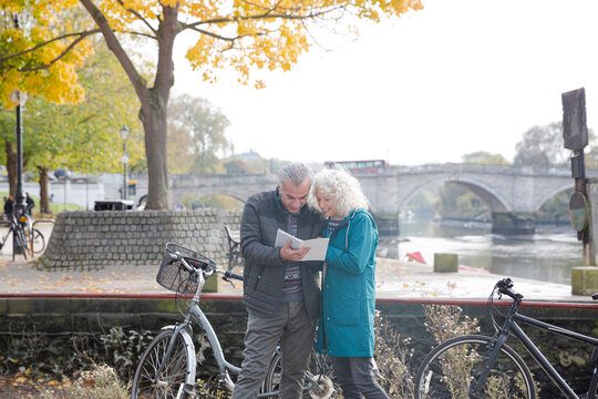 Senior couple with bicycles traveling, looking at guidebook along autumn river - Powered by Adobe