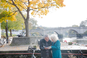 Senior couple with bicycles traveling, looking at guidebook along autumn river