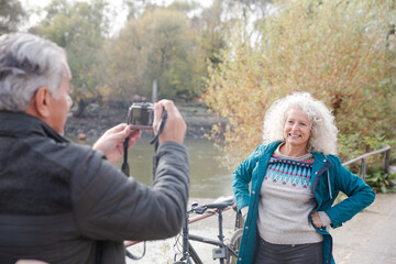 Senior man photographing wife with bicycle in autumn city