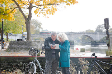 Senior couple with bicycles traveling, looking at guidebook along autumn river