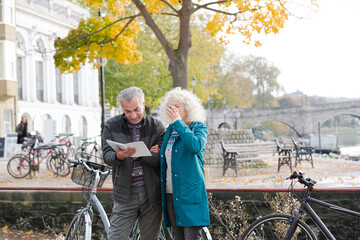 Senior couple with bicycles traveling, looking at guidebook along autumn river