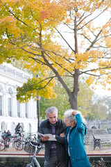 Senior couple with bicycles traveling, looking at guidebook along autumn river