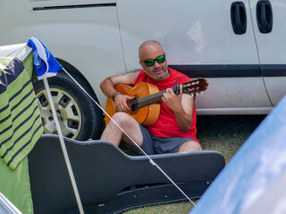 A traveler in a red shirt, with sunglasses and shaved hair, plays the Spanish guitar sitting on the...