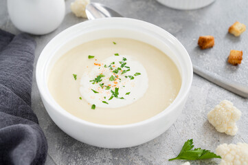 Homemade cauliflower soup puree in a white bowl with cheese, cream, spices and fresh parsley on a gray concrete background. Top view, copy space.