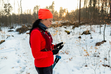 Female Runner Checking Mobile While Running in Woods. Sportswoman With Trekking Poles Holding Cellphone in Hands On Cold Winter Day. - obrazy, fototapety, plakaty