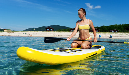 Tanned woman in bikini sitting on surfboard with paddle relaxing at sea resort enjoying summer