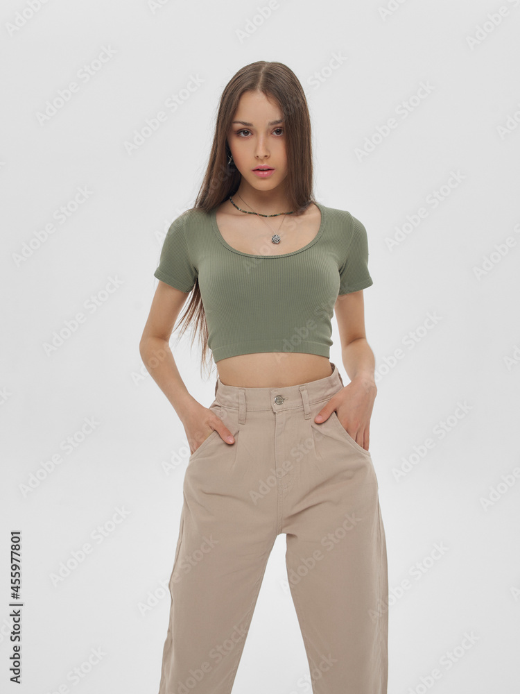 Wall mural Young elegant woman with long straight hair and natural make-up in green top and beige jeans standing and posing on bright grey background