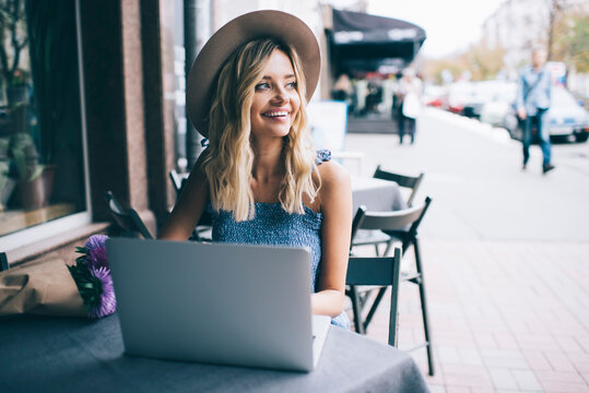 Cheerful female digital nomad with modern laptop computer working remotely in street cafe enjoying freelance lifestyle, happy Caucasian hipster girl in stylish hat using netbook and 4g wireless