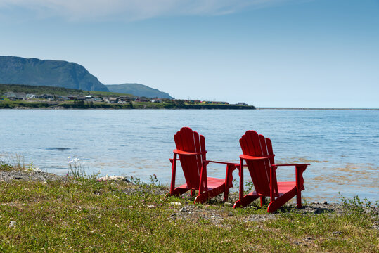 Two bright empty red Adirondack chairs on the edge of a green grassy meadow overlooking the blue ocean with mountains covered in trees, a bay, and cove. The water is calm and the sky is blue. 
