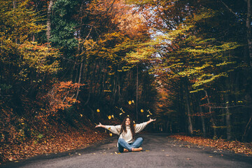 Positive curly haired woman in sweater throwing dry leaves in picturesque autumn forest with colorful trees on sunny day.