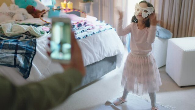 happy little girl playing dress up wearing mask in bedroom with mother taking photo using smartphone sharing on social media