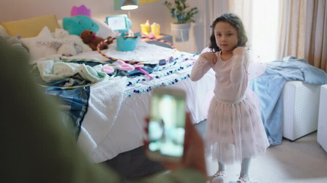 happy little girl playing dress up wearing fairy wings in bedroom with mother taking photo using smartphone sharing on social media