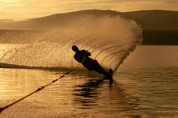 Silhouette of a Man practicing water ski slalom at a lake in the sunrise sunset making water spray...