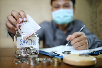 An Asian businessman in a gray shirt is wearing a face mask and is dropping his banknotes to save money. Financial and accounting concept.