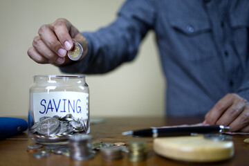 A gray-shirted Asian businessman is dropping coins into a glass jar for savings. Financial and accounting concept.
