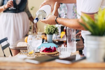 Hands of man pouring alcohol into glass preparing cocktail or a refreshing drink on lunch table while woman in drinking in the background. Preparing drinks for the celebration