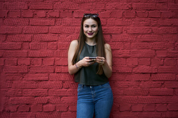 Cheerful Caucasian blogger with modern cellular gadget for networking influence content standing at publicity area with copy space for advertising text, smiling hipster girl with digital smartphone