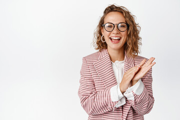 Business people concept. Young modern female manager in glasses and suit, applause and smile pleased, praise you with clap gesture, standing over white background