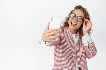 Happy business woman in glasses and suit taking selfie, laughing and smiling, using photo filter app on mobile phone, white background