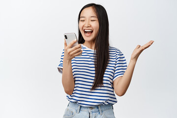 Image of asian brunette girl looking with excited and surprised face at smartphone, reacting amazed...