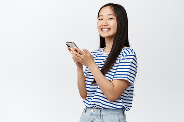 Portrait of asian girl using smartphone and smiling at camera, concept of cellular technology and mobile applications, white background
