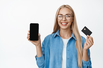 Beautiful blond girl in glasses smiling, showing mobile phone screen and credit card, demonstrating finance app, standing over white background