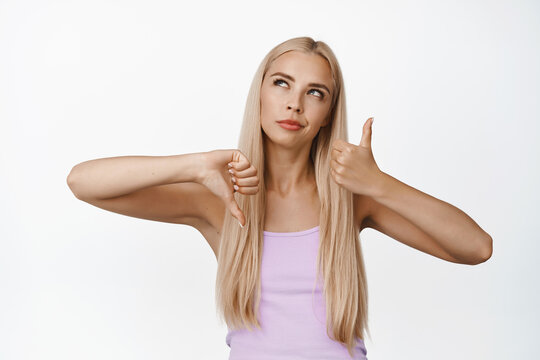 Thoughtful Blond Girl Shows Thumbs Up And Down, Making Decision, Weighing Pros And Cons, Like Or Dislike, Stands Over White Background