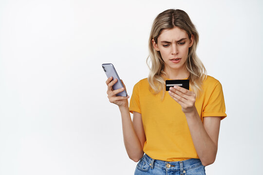 Image of serious blond girl holding mobile phone, looking at credit card number with concentrated face, trying to pay online, standing over white background