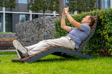 Millennial man photographing himself. Handsome man relaxing on wooden chaise lounge and taking ...