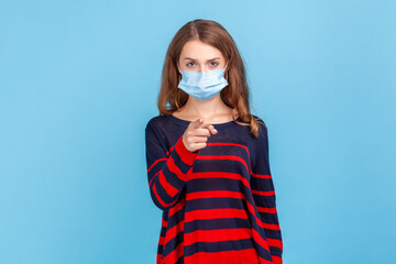 Hey you. take care of you health. Portrait of serious woman wearing sweater and surgical medical mask standing, pointing and looking at camera and blame. Indoor studio shot isolated on blue background