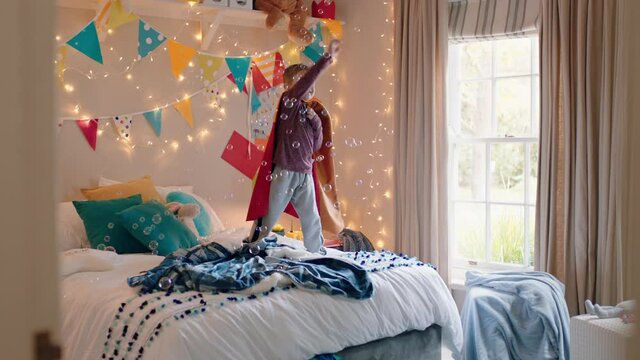 happy little boy jumping on bed wearing costume playing game with toy gun shooting bubbles enjoying playful imagination in colorful bedroom at home