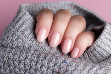 Female hand in gray knitted sweater with beautiful manicure - pink nude nails on pink background. Nail care concept