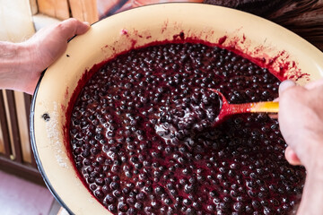Hands stir with a wooden spoon jam from blueberries in a metal basin. Traditional preparation of a sweet dessert in the countryside. 