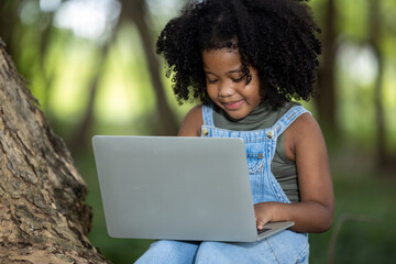 Obraz na płótnie Canvas children girl African American ethnicity black skin sitting on tree base use Laptop computer to chat with friends via video call with 5G internet signal in the park