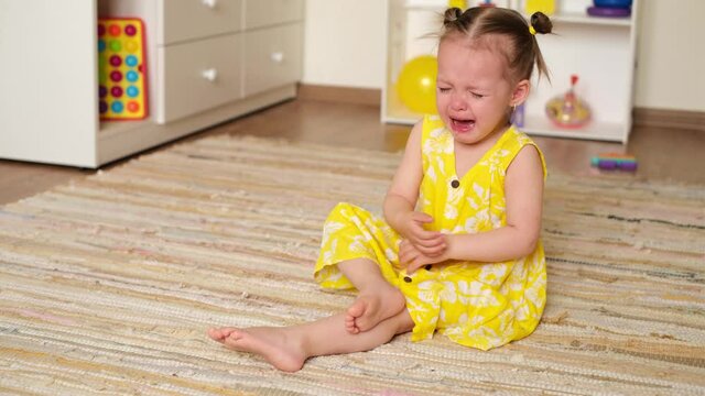 A small girl is sitting on the floor of the nursery and crying. Upbringing and psychology of children. The crisis of a two-year-old kid is hysterics and whims. Capricious baby